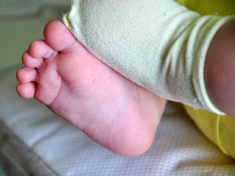 Foto dos pés de um bebê recém-nascido. Um pé está descalço e o outro está coberto por uma meia na cor verde claro. Ao fundo, uma pequena cama de hospital está desfocada, sugerindo que a foto foi captada em uma maternidade. No canto da imagem aparece uma parte da roupa do bebê na cor amarela